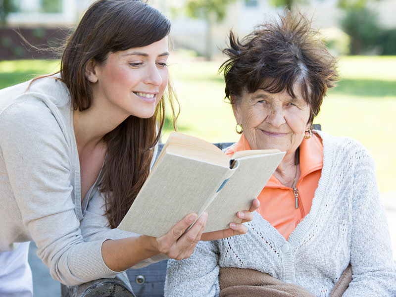 Eine junge Frau liest einer Seniorin im Freien aus einem Buch vor.